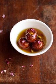gulab jamun with milk powder garnished with dried rose petals and pistachios and served in a white bowl.