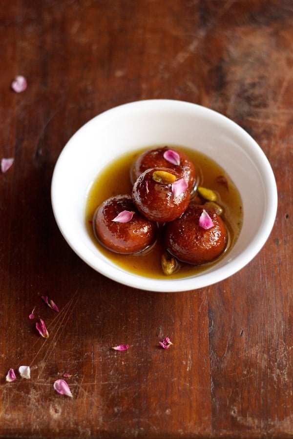 gulab jamun with milk powder garnished with dried rose petals and pistachios and served in a white bowl.