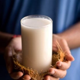 coconut milk served in a glass inside an empty coconut shell being held by hands.