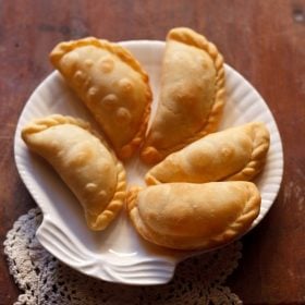 five karanji pieces on a white plate on a cream doily