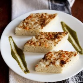 white dhokla cut into diamonds and served with coriander chutney on a white plate.