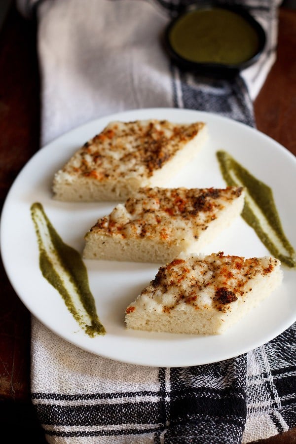 white dhokla cut into diamonds and served with coriander chutney on a white plate.