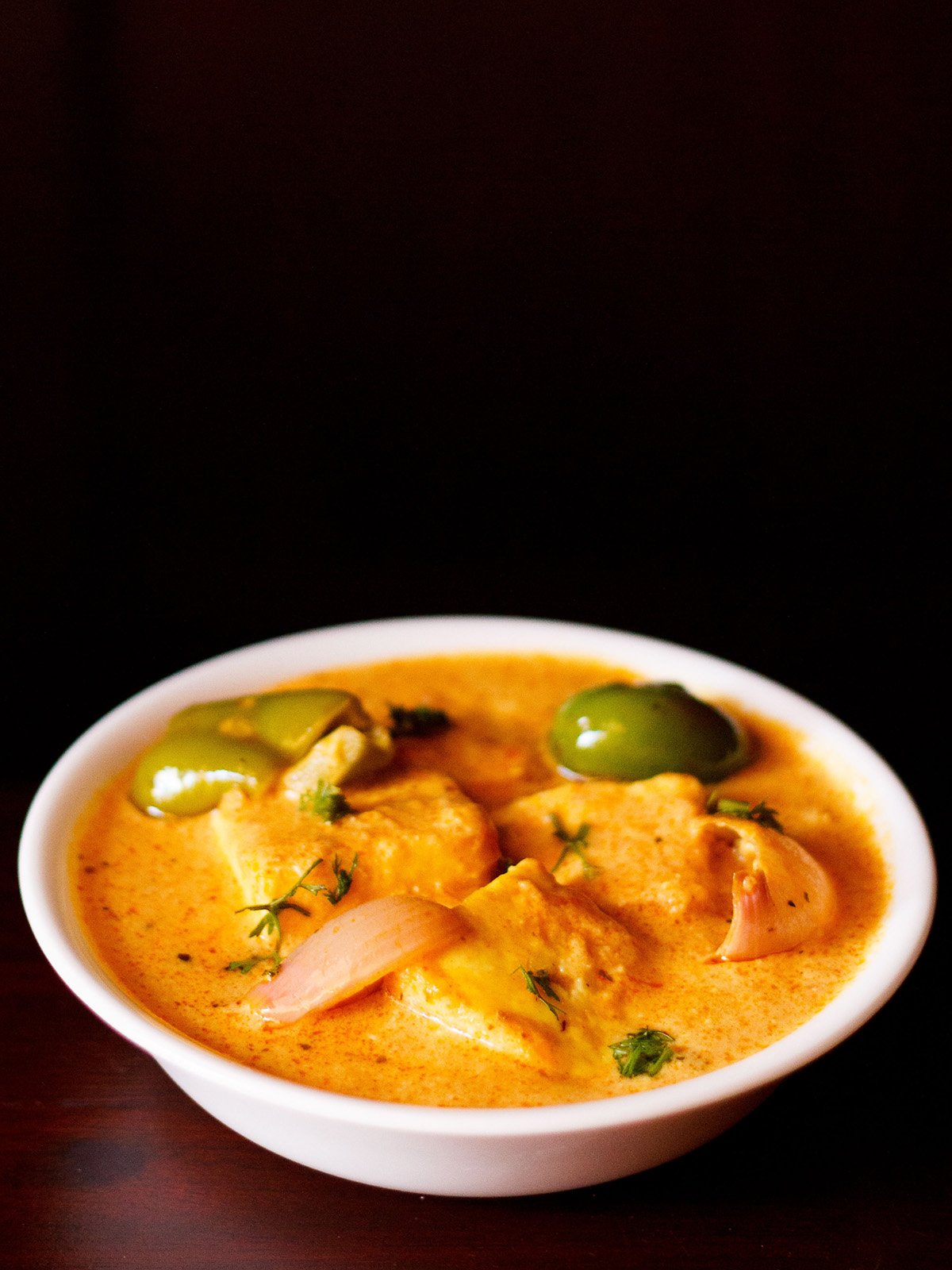 paneer tikka masala served in a white bowl garnished with some coriander leaves on a dark mahogany board.