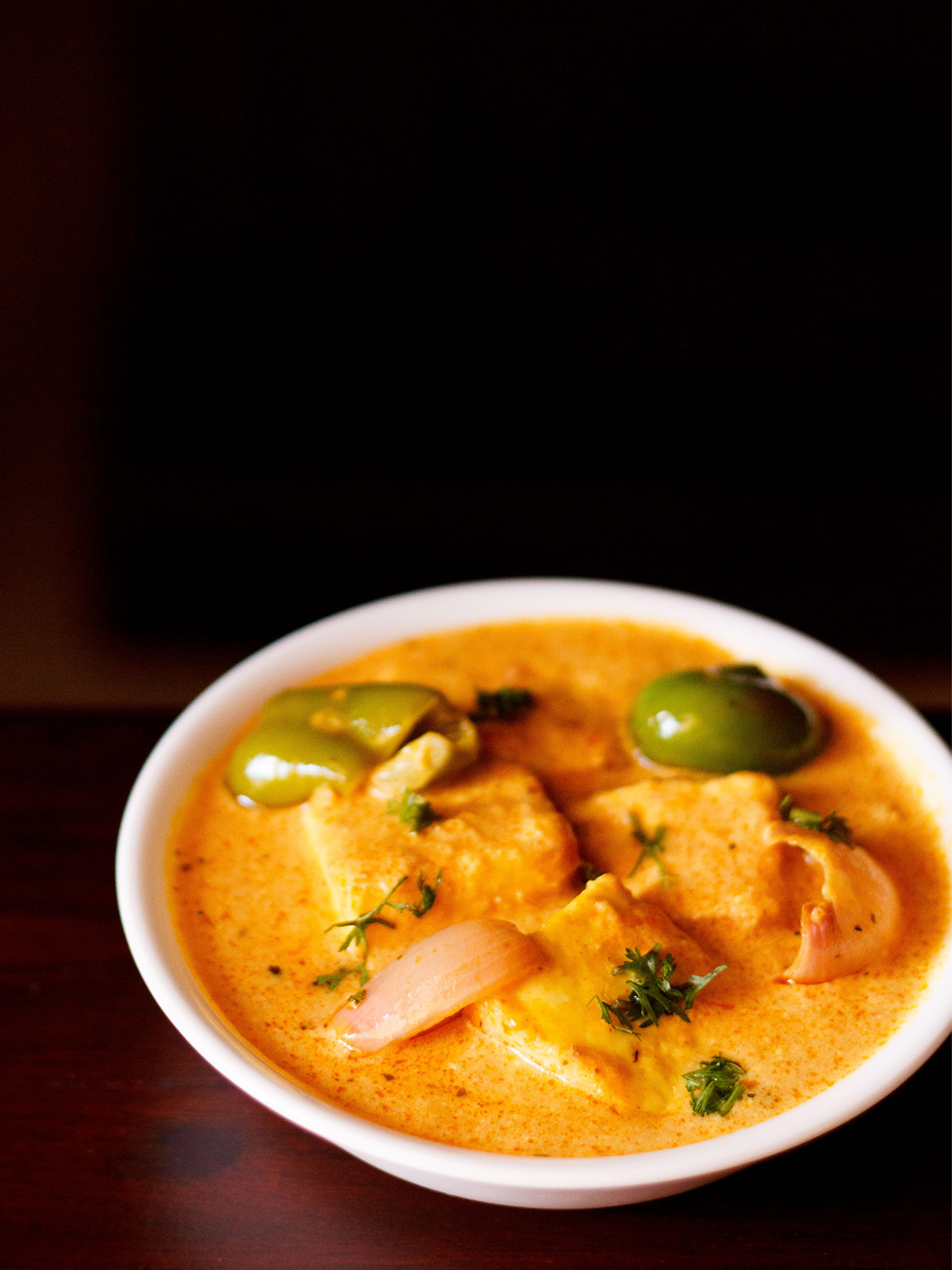 paneer tikka masala served in a white bowl garnished with some coriander leaves on a dark mahogany table 