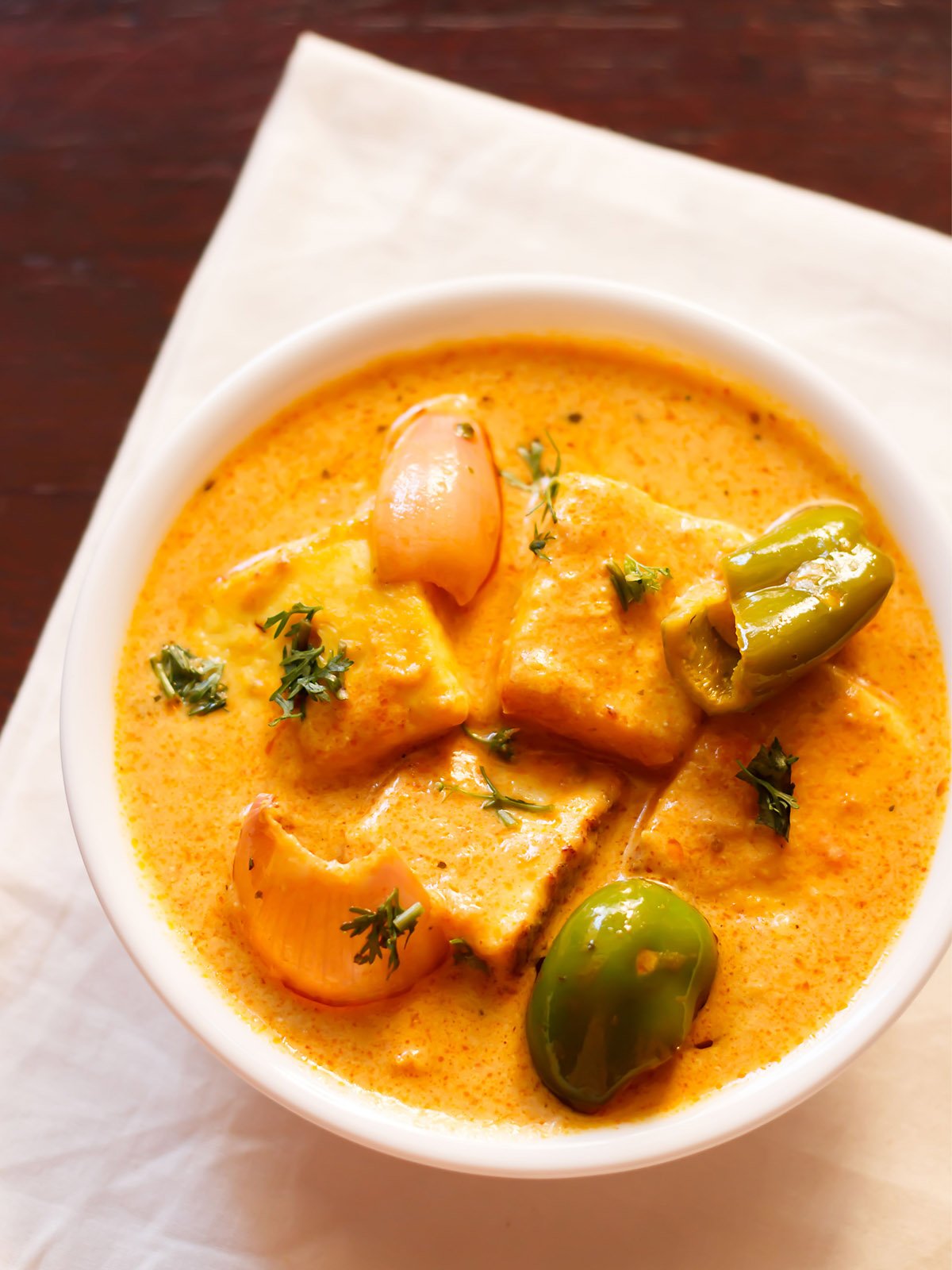 paneer tikka masala adornado con unas hojas de cilantro en un bol blanco colocado sobre una servilleta blanca en una mesa de caoba oscura