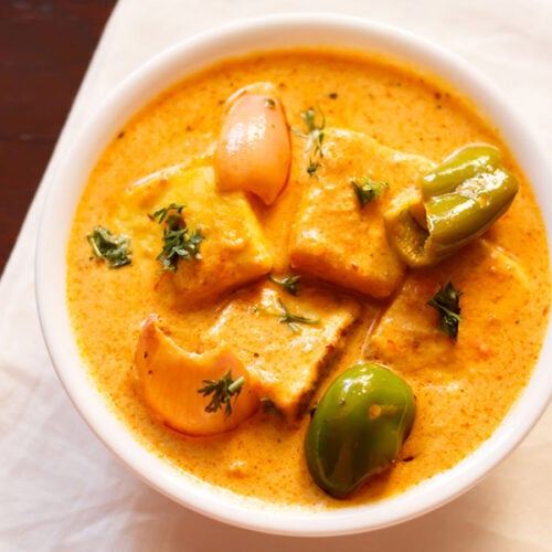 paneer tikka masala podany w białej misce przyozdobionej kilkoma liśćmi kolendry na ciemnym mahoniowym stole