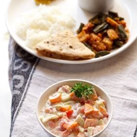 veg raita garnished with a coriander leaf and served in a bowl with a plate of Indian meal kept in the background.