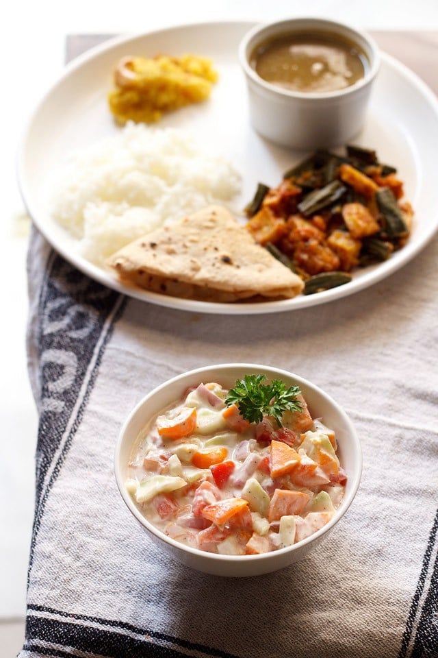 veg raita garnished with a coriander leaf and served in a bowl with a plate of Indian meal kept in the background.