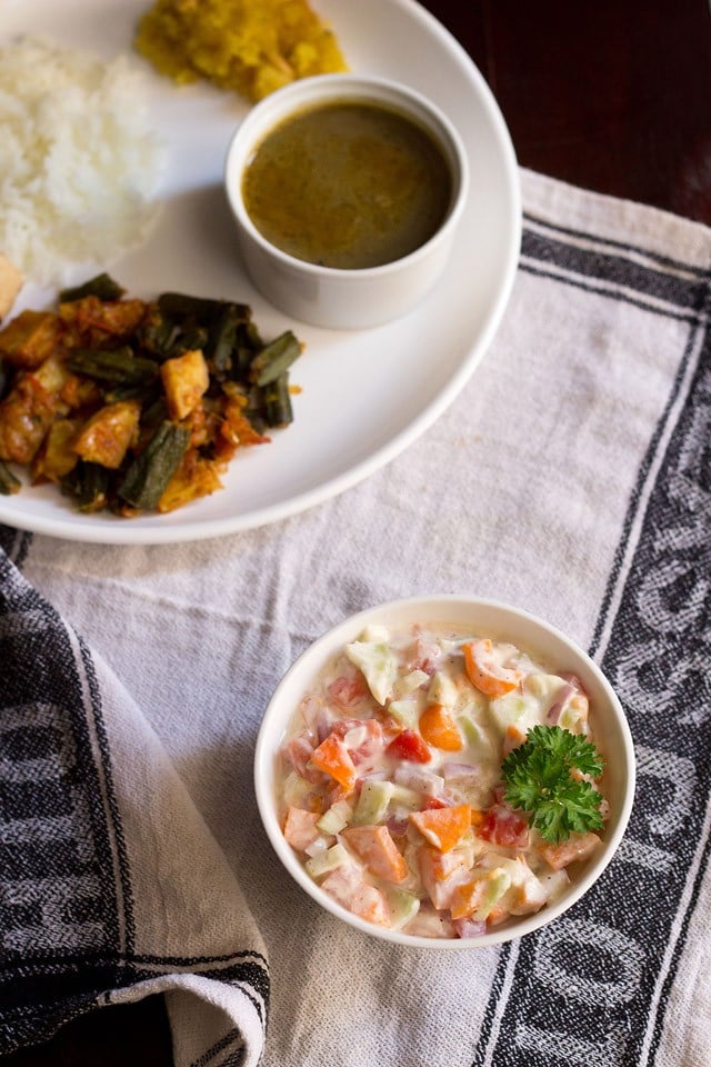 veg raita garnished with a coriander leaf and served in a bowl with a plate of Indian meal kept in the background. 