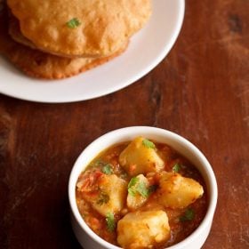 aloo tamatar ki sabji served in a white bowl with pooris on a white plate kept on the top left side.
