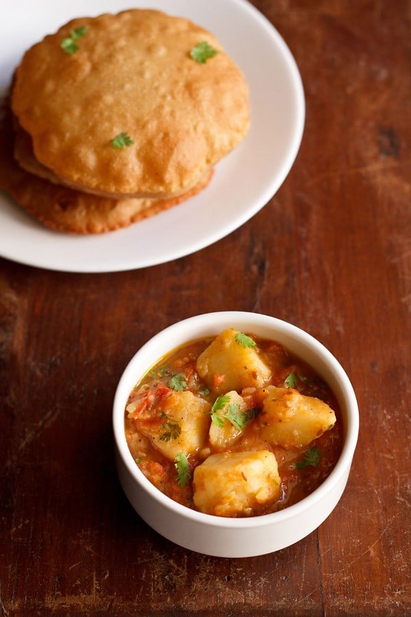 aloo tamatar ki sabji served in a white bowl with pooris on a white plate kept on the top left side.