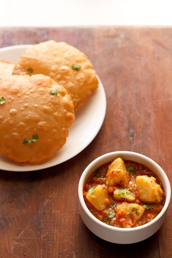 aloo tamatar ki sabji served in a white bowl with pooris on a white plate kept on the top left side.