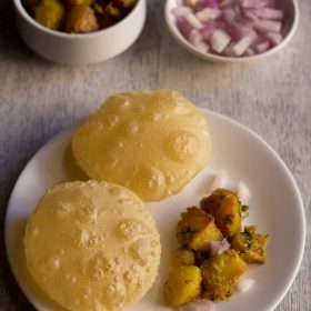 luchi served on a white plate with aloo sabzi and a bowl of sabzi and a bowl of chopped onions kept on the upper side.