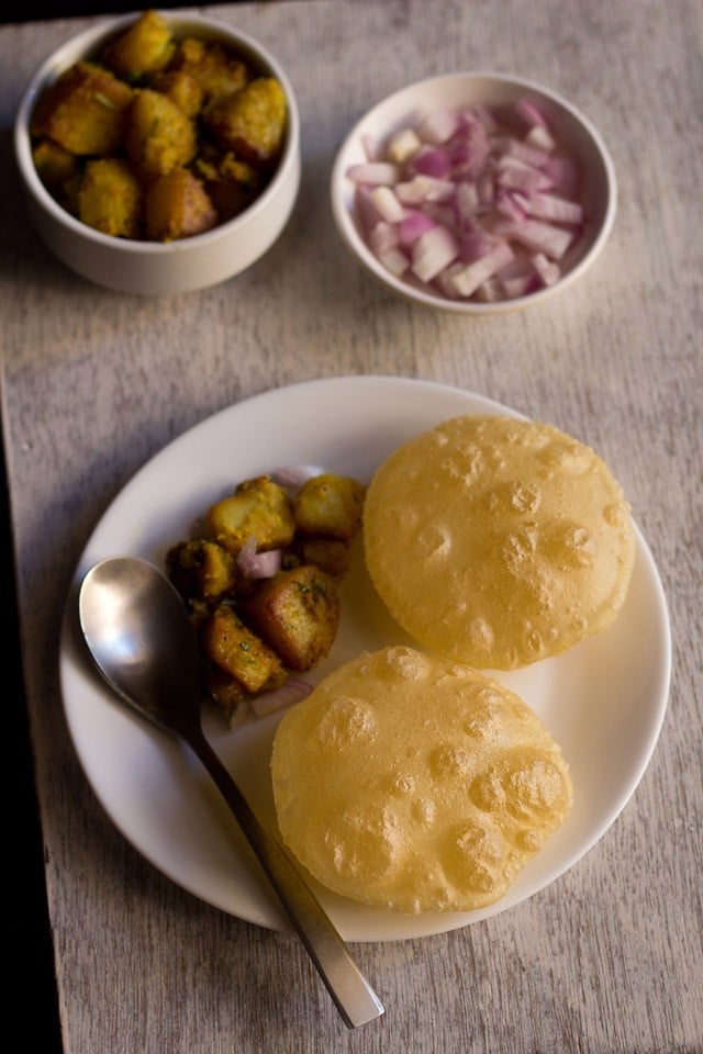 luchi served on a white plate with aloo sabzi, a spoon and a bowl of sabzi and a bowl of chopped onions kept on the upper side. 