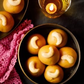 besan ke laddu placeret i en messingbeholder med en mørk lyserød doily ved siden og et glas diya på toppen