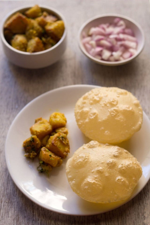 luchi served on a white plate with aloo sabzi and a bowl of sabzi and a bowl of chopped onions kept on the upper side. 