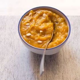 pumpkin halwa garnished with melon seeds and served in a blue rimmed white bowl with a spoon inside it.