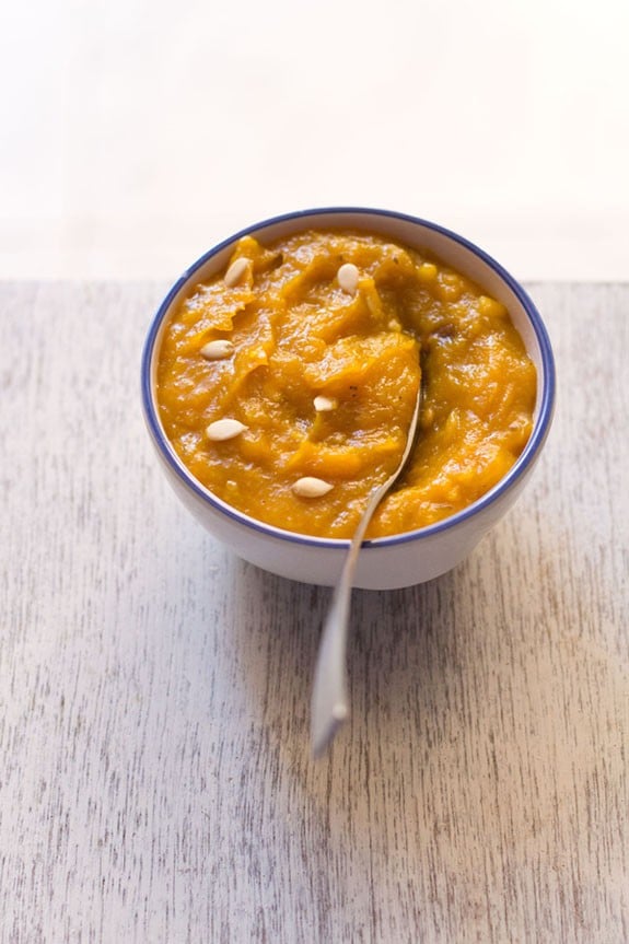 pumpkin halwa garnished with melon seeds and served in a blue rimmed white bowl with a spoon inside it.