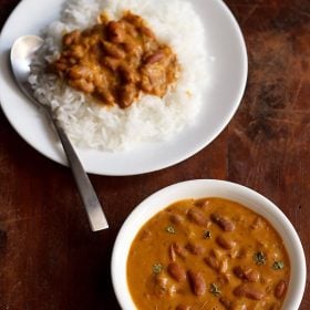rajma masala garnished with a few kasuri methi and served in a white bowl on a brown wooden board and a plate of rice and rajma curry with a spoon kept on left top side of the board