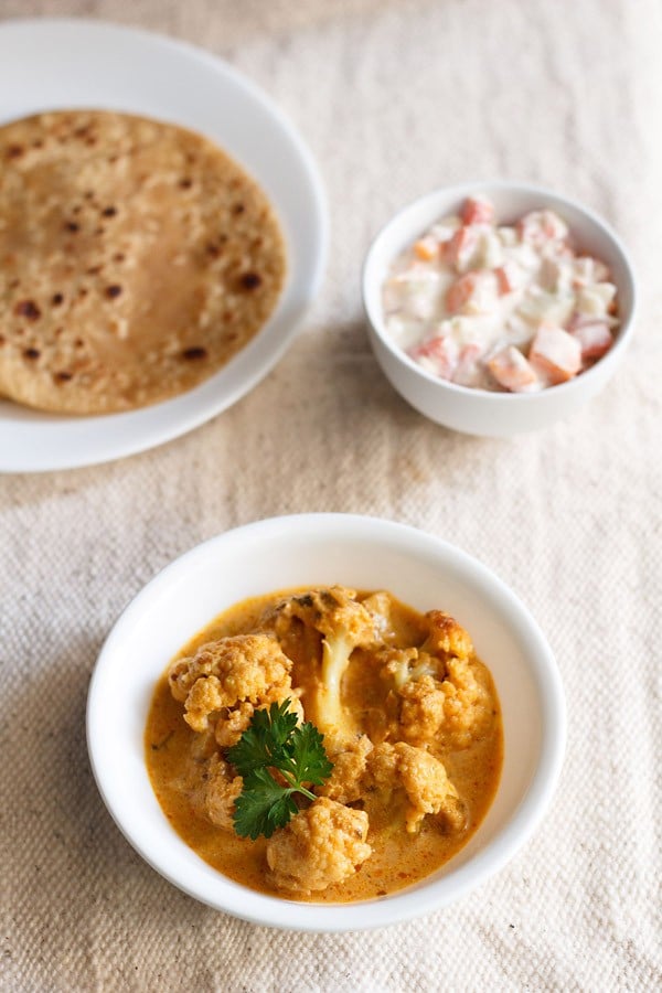 gobi masala garnished with coriander leaf and served in a bowl with a plate of chapattis kept on the left side and a small bowl of raita kept on the right side. 