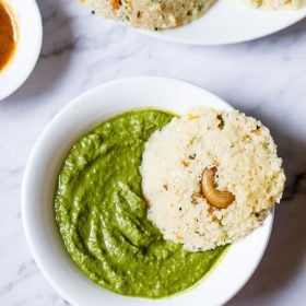 A single Rava Idli dipped partly in a bowl of coriander chutney (cilantro dip) on a white marble backdrop