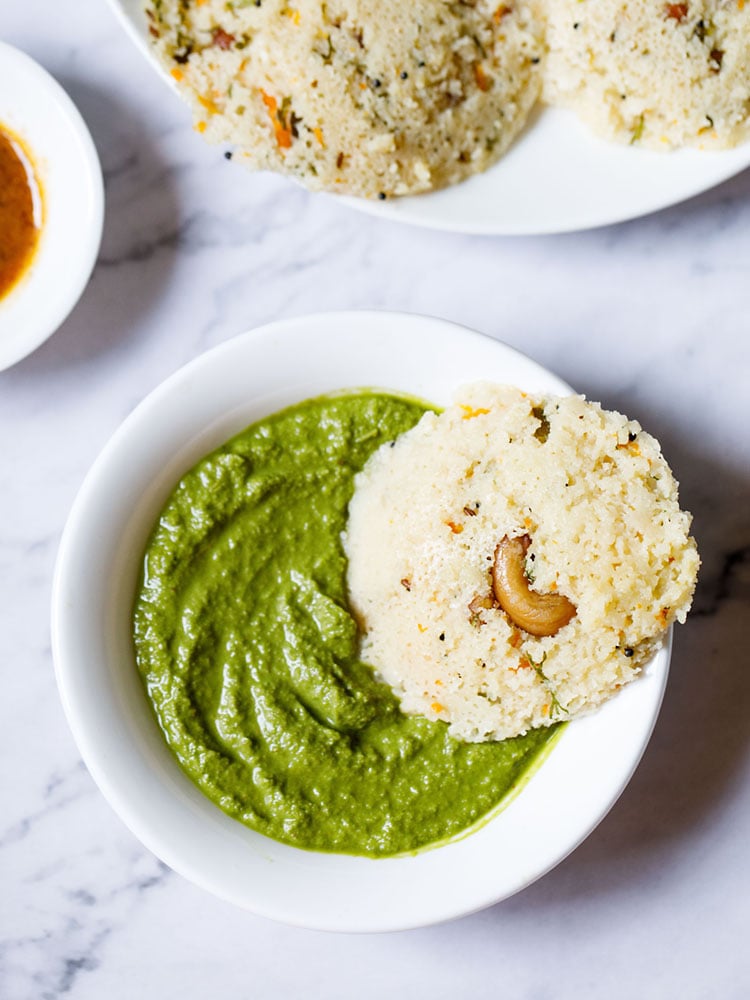 A single Rava Idli dipped partly in a bowl of coriander chutney (cilantro dip) on a white marble backdrop