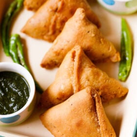 samosa arranged in a line on a cream tray with chutneys in small bowls and fried green chillies on the tray