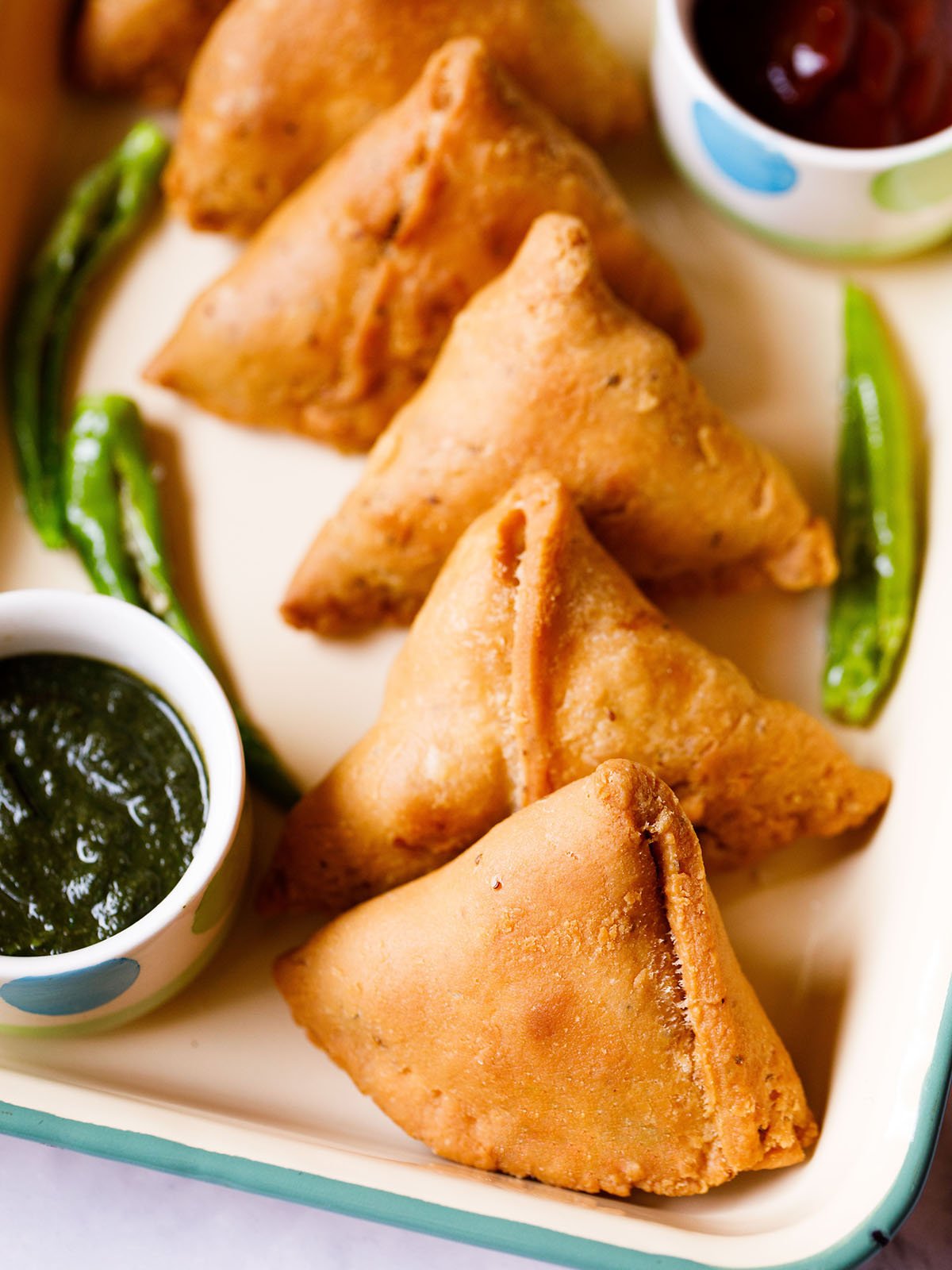 samosa arranged in a line on a cream tray with chutneys in small bowls and fried green chillies on the tray