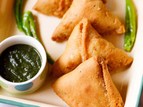 samosa kept in a cream tray with a bowl of green chutney and fried green chillies by the side