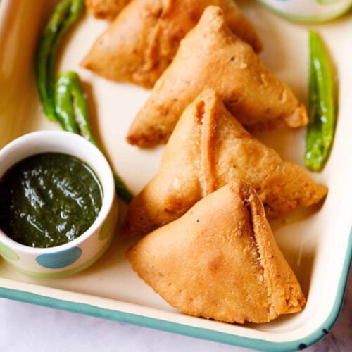 samosa kept in a cream tray with a bowl of green chutney and fried green chillies by the side