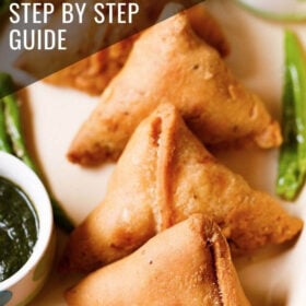 samosa kept in a cream tray with a bowl of green chutney and fried green chillies by the side