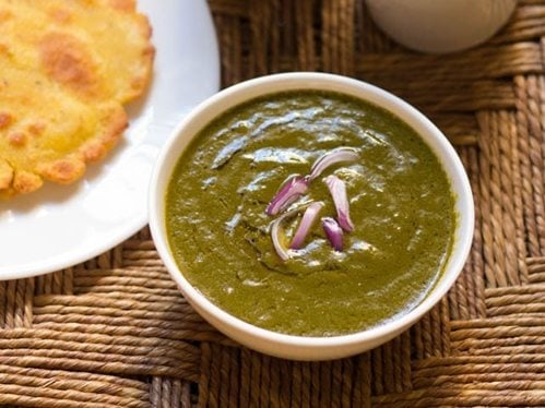 sarson ka saag in a white bowl on a rattan table with a white plate of makki ki roti to the side.