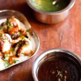 tamarind chutney garnished with one sprig of coriander served in a steel bowl with a square plate of dahi vada kept in the left center and green chutney kept in a steel bowl on top.