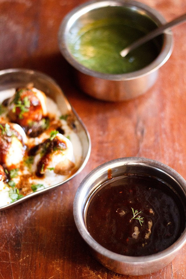 tamarind chutney garnished with one sprig of coriander served in a steel bowl with a square plate of dahi vada kept in the left center and green chutney kept in a steel bowl on top.