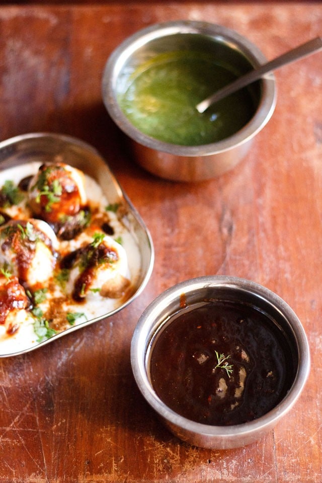 tamarind chutney garnished with one sprig of coriander served in a steel bowl with a square plate of Dahi Vada kept in the left center and green chutney kept in  a steel bowl on top