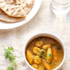 baby corn masala garnished with a coriander sprig and served in a white bowl with a plate of chapatis kept in the background.