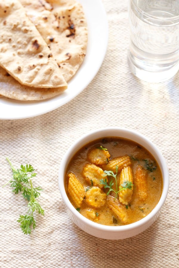 baby corn masala garnished with a coriander sprig and served in a white bowl with a plate of chapatis kept in the background.