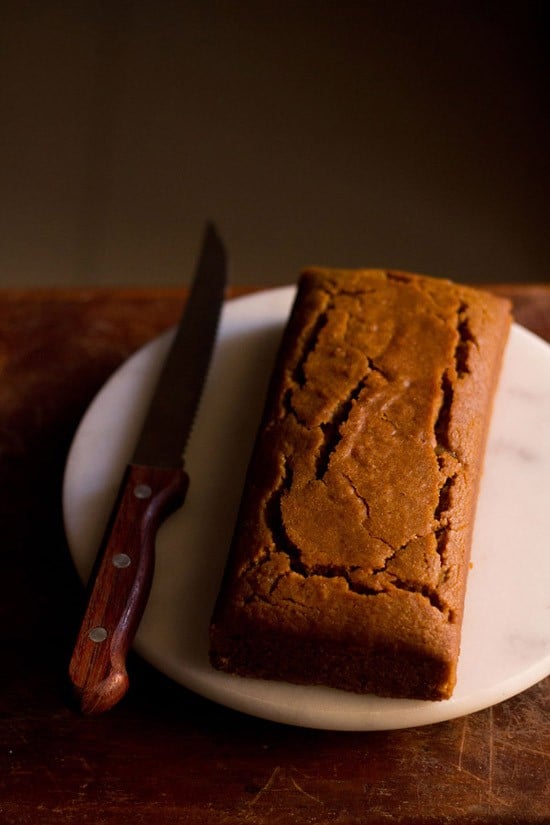 eggless gingerbread cake in a loaf shape on a round marble cutting board with a serrated knife to the side.