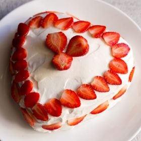 दिल के आकार का स्ट्रॉबेरी केक व्हीप्ड क्रीम से फ्रॉस्ट किया गया और एक सफेद प्लेट पर स्ट्रॉबेरी से सजाया गया