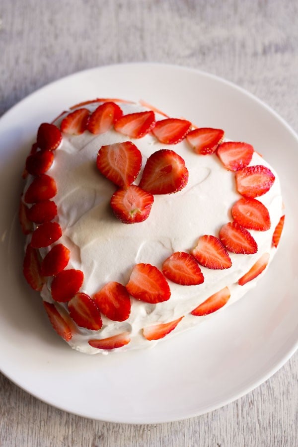 heart-shaped strawberry cake frosted with whipped cream and decorated with strawberries on a white plate