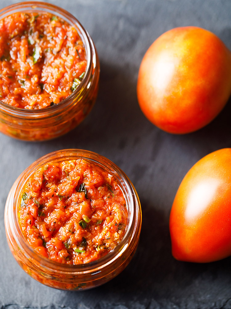 pizza sauce in two small glass jar placed on a dark gray slate board with fresh two tomatoes by the side