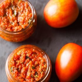 pizza sauce in two small glass jar placed on a dark gray slate board with fresh two tomatoes by the side