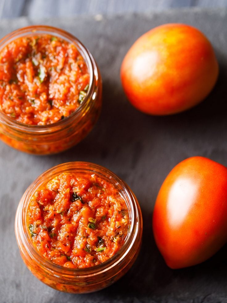 pizza sauce in two small glass jar placed on a dark gray slate board with fresh two tomatoes by the side.