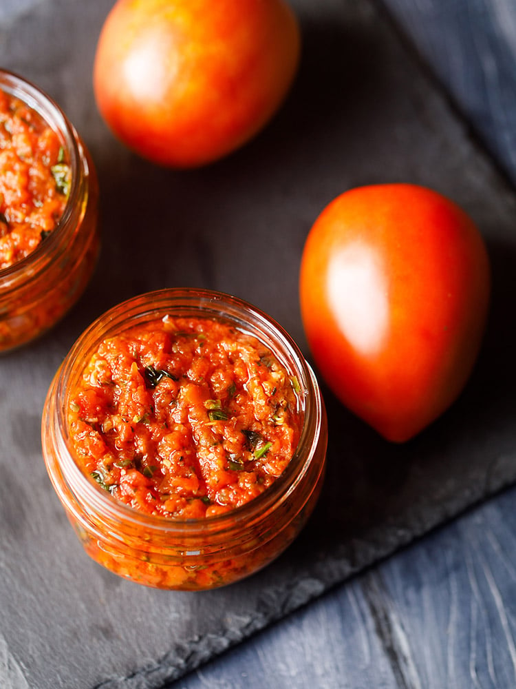 pizza sauce in two small glass jar placed on a dark gray slate board with fresh two tomatoes by the side