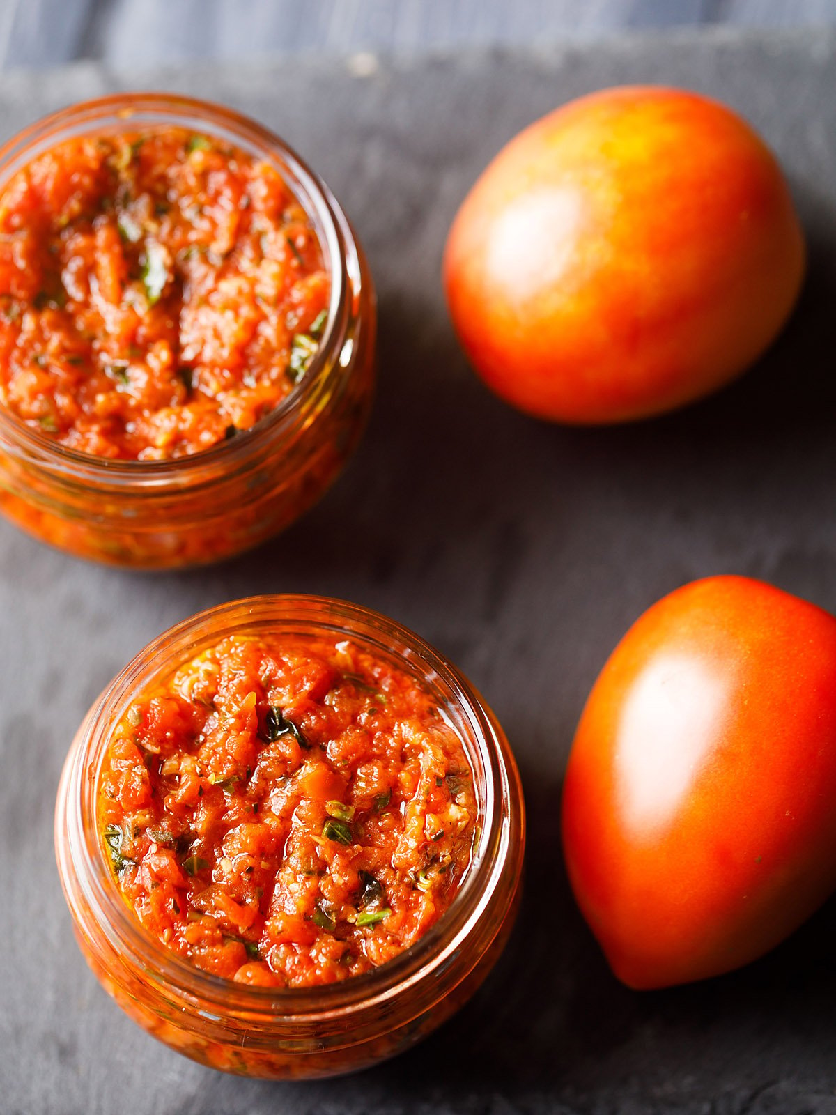 pizza sauce in two small glass jar placed on a dark gray slate board with fresh two tomatoes by the side