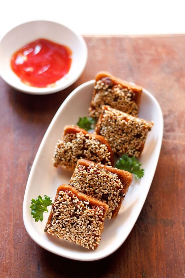 sesame toast garnished with coriander leaves and served on a white platter with red chili sauce.