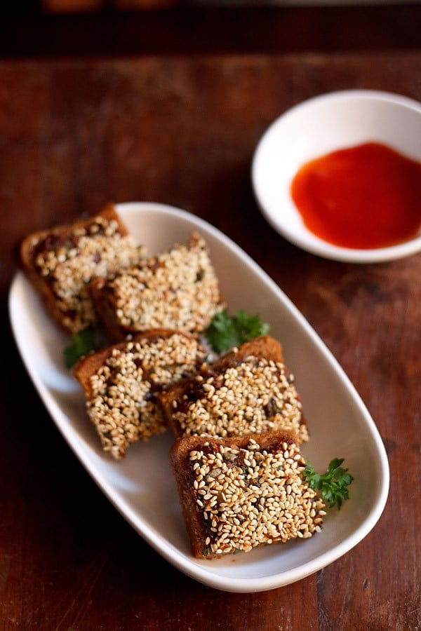 sesame toast garnished with coriander leaves and served on a white platter with red chili sauce. 