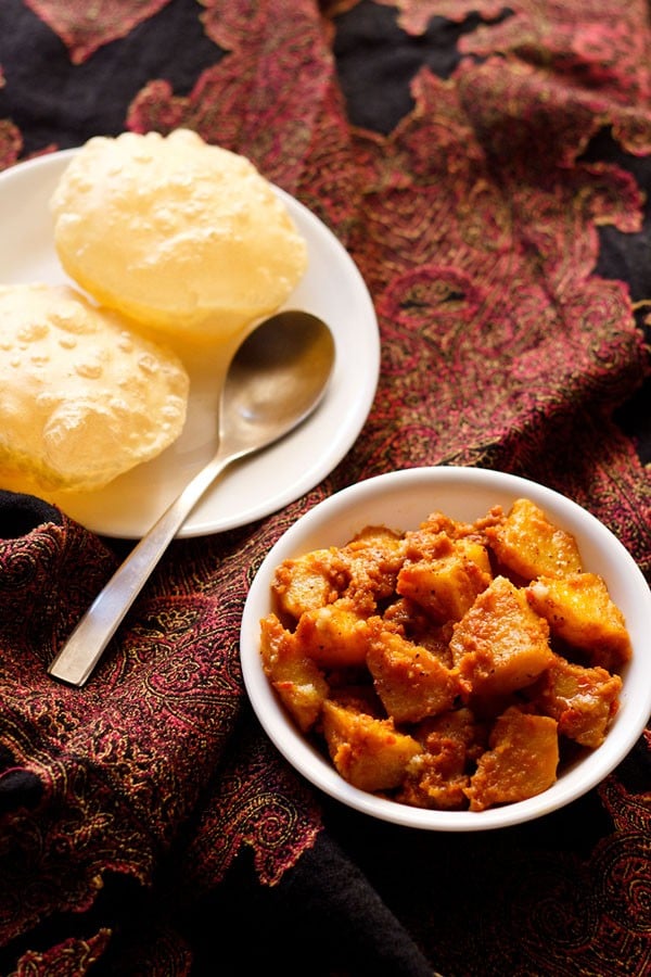 alur dom served in a white bowl with luchi on a white plate and a spoon kept on the side.