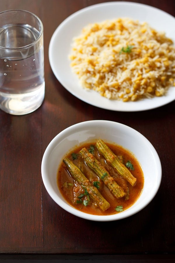 drumstick curry garnished with chopped coriander leaves and served in a white bowl with a plate of dal pulao and a glass of water kept in the background.