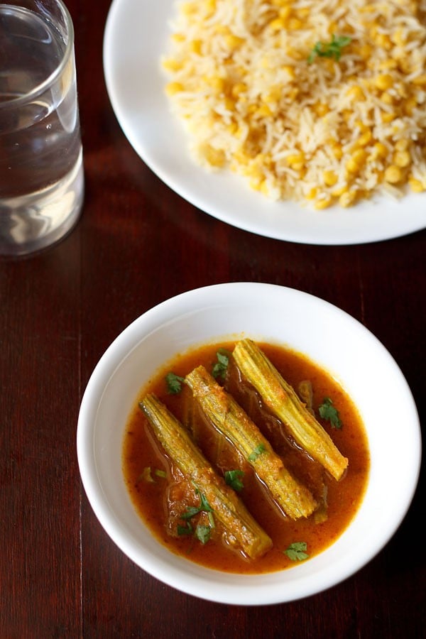 drumstick curry garnished with chopped coriander leaves and served in a white bowl with a plate of dal pulao and a glass of water kept in the background. 